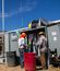 Two workers wearing hard hats and protective gear stand beside an open electrical cabinet at an outdoor industrial jobsite. They handle cables and components while performing electrical work.