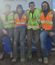 Four construction workers sit close together on a blue equipment box, smiling at the camera. They are wearing worn, muddy work boots, hoodies, and brightly colored safety vests—two in orange and two in yellow.