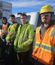 Four construction workers stand side by side in front of a white work truck on a sunny day. They wear hoodies, worn work pants, and high-visibility safety vests in orange and yellow. The worker on the far right also wears a yellow hard hat and safety glasses.