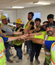 A group of construction team members stand in a circle inside a breakroom or jobsite trailer, all extending one hand toward the center in a team huddle gesture. They are smiling and wearing high-visibility vests, hard hats, and work gear.