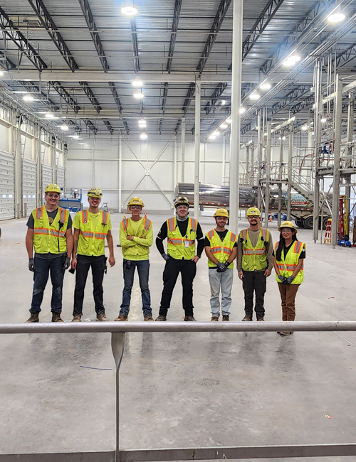 A line of seven interns wearing personal protective equipment including helmets, vests, and safety glasses, standing in a large industrial facility with metallic structures around.
