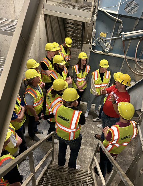 A group of interns, equipped with safety gear, listening to a supervisor explain machinery on a job site