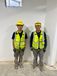 Two Interstates workers standing side by side in a construction area, wearing yellow hard hats, safety glasses, high-visibility vests, gloves, and work boots in front of a white wall.