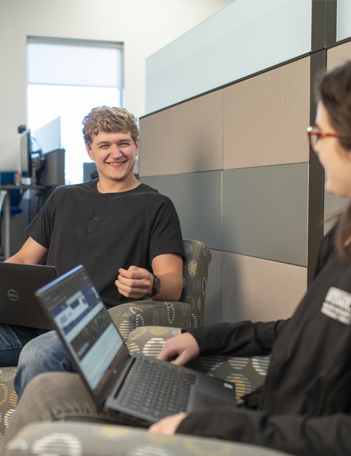 A young intern laughs during a discussion with a coworker at a laptop, both seated in a modern office space, showcasing a relaxed and friendly work environment.