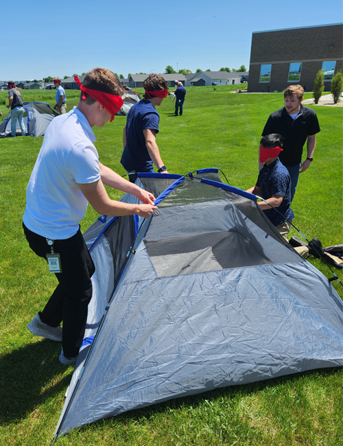 Interns participating in an outdoor team-building activity, working together to assemble a tent while blindfolded, under sunny skies.