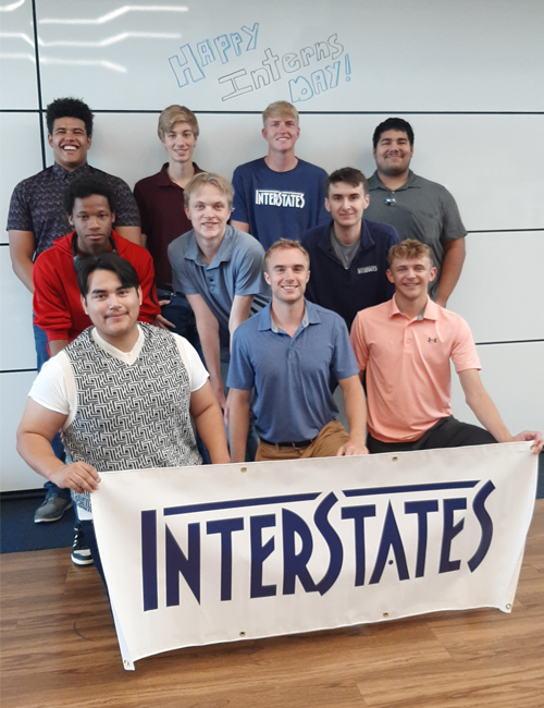 Interns holding a banner that reads 'INTERSTATES,' celebrating Interns Day, posing happily in a modern office environment with "Happy Intern's Day!" written on the whiteboard behind them.