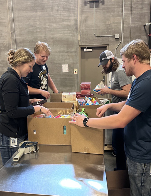 Interns volunteering by packing assorted food items into large boxes in a warehouse, engaged in teamwork and community service.