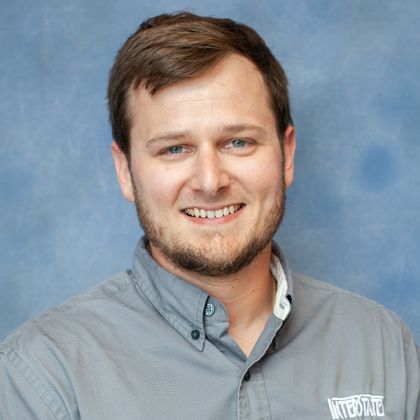 Headshot of a smiling man with short brown hair and a beard wearing a gray button-up shirt.