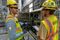 Two workers in hard hats and safety vests talk beside an open electrical control panel in an industrial facility.