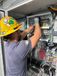 An electrician wearing a yellow hard hat and gloves is wiring the inside of an electrical control panel. Colored cables and labeled components are organized across the panel, with a warning label visible below.