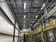 High‑bay industrial room with exposed pipes and conduit under bright strip lights; a yellow‑railed catwalk and stairs lead to a platform with large fans at the far end.