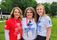 Three women wearing Midwest Honor Flight T-shirts stand together outdoors on a grassy lawn, smiling at the camera.
