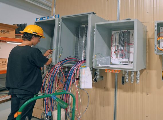 **Accessibility text (alt text):**  An electrical technician wearing a yellow hard hat wires open electrical control panels inside a manufacturing facility. Several gray enclosures are mounted along a wall, with organized wiring, conduit, and cable bundles visible as the technician assembles and connects the equipment.