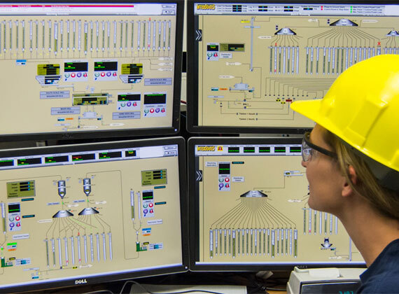 A worker wearing a yellow hard hat and safety glasses monitors multiple screens displaying a complex industrial control system interface.