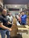 People in a warehouse stand beside a long row of open cardboard boxes, smiling and handling packing materials.