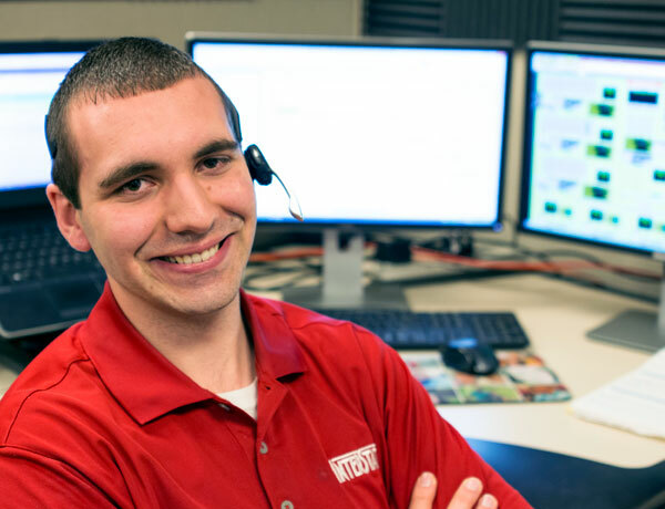 Support employee smiles at the camera, several monitors are in the background.