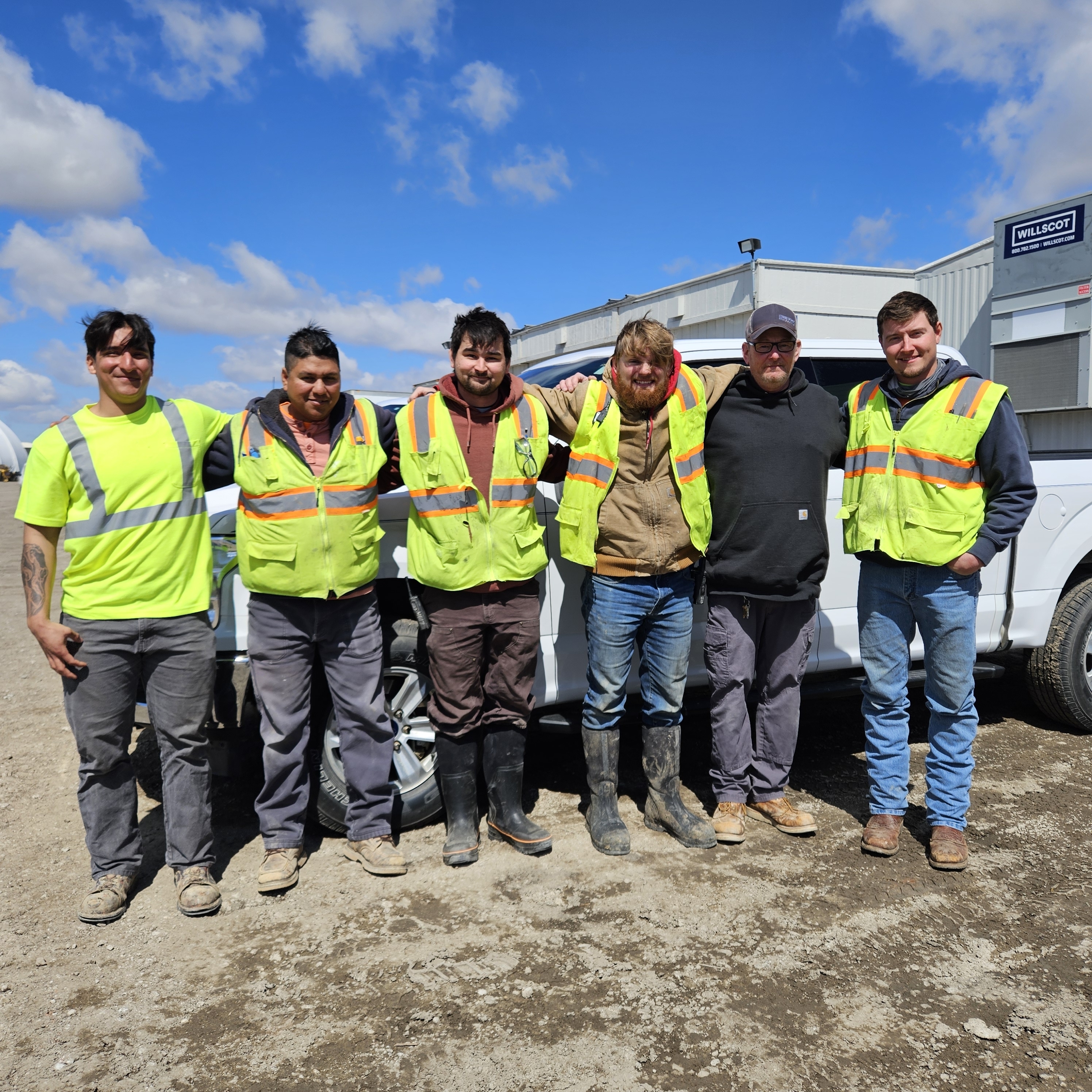 Six field workers in safety vests and work gear lined up against company vehicles, displaying teamwork in industrial setting