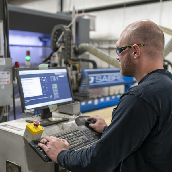 A technician wearing safety glasses stands at a computer workstation, using software to control a CNC machine in an industrial workshop setting.