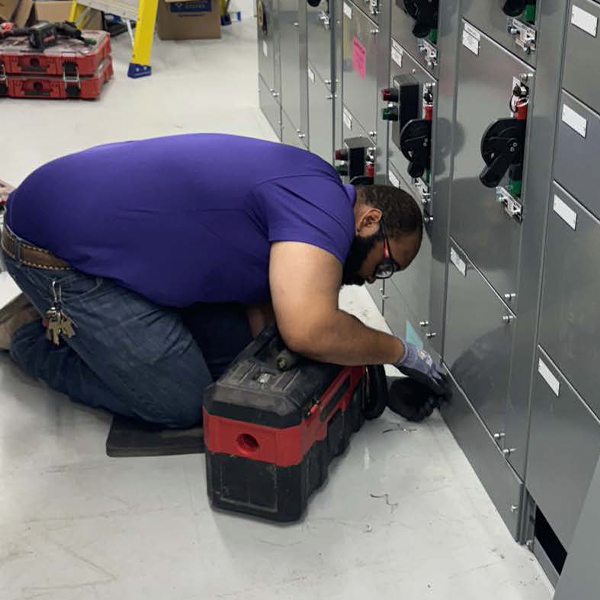 A technician in a purple shirt kneels on the floor in front of a row of gray electrical switchgear panels, using tools from a red and black toolbox to work on the lower section.