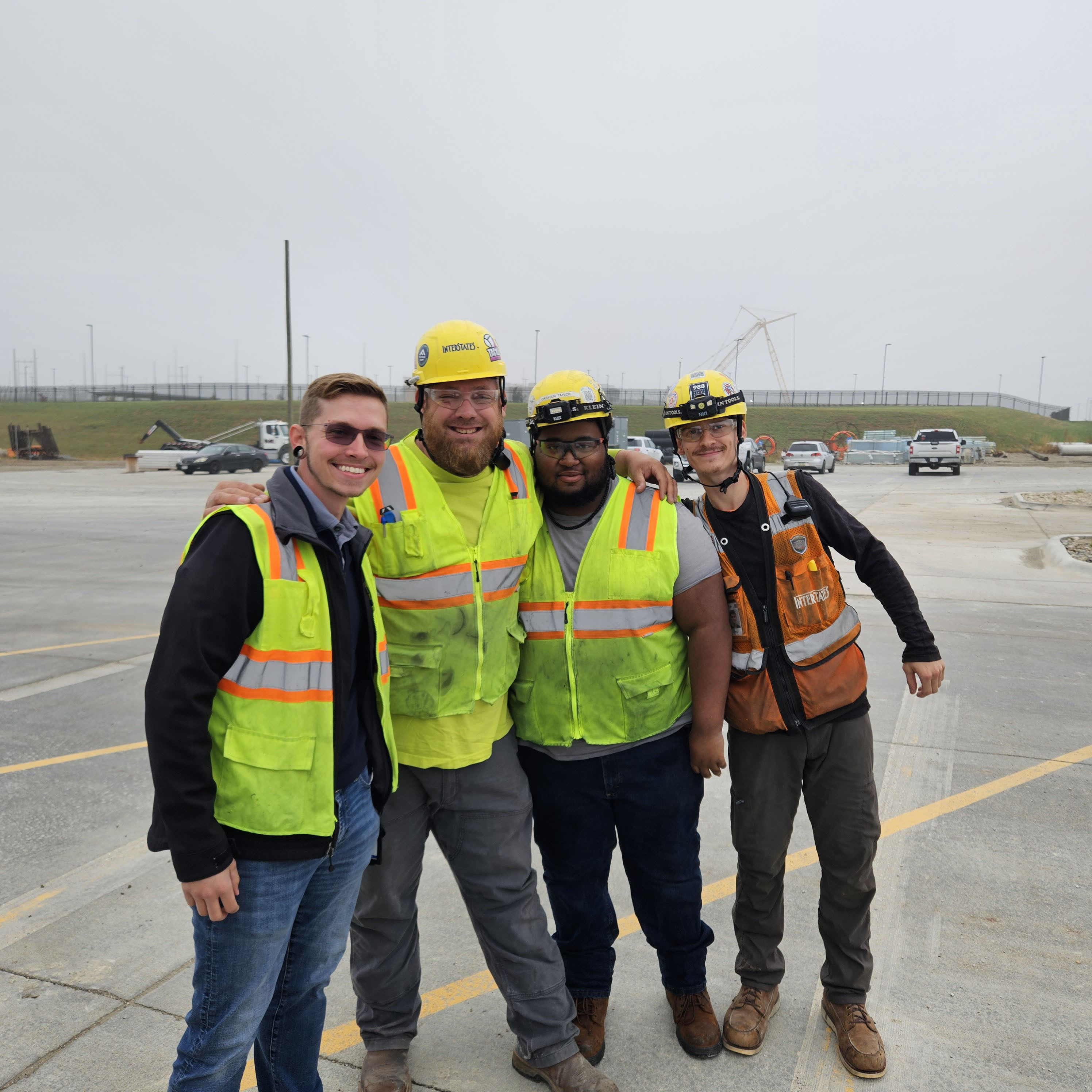 Four field team members in hard hats and safety vests posing together on construction site, with power lines visible in background