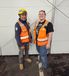 Two workers in safety gear - one wearing a yellow hard hat and navy shirt, the other in a black t-shirt, both wearing orange reflective vests - standing against a white wall in what appears to be an industrial setting