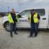 Two men wearing high-visibility yellow jackets stand in front of a white Interstates pickup truck on a gravel jobsite. Both are smiling toward the camera.