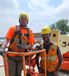Two people in safety vests, safety hats, safety glasses, and one tied in a lift smiling.