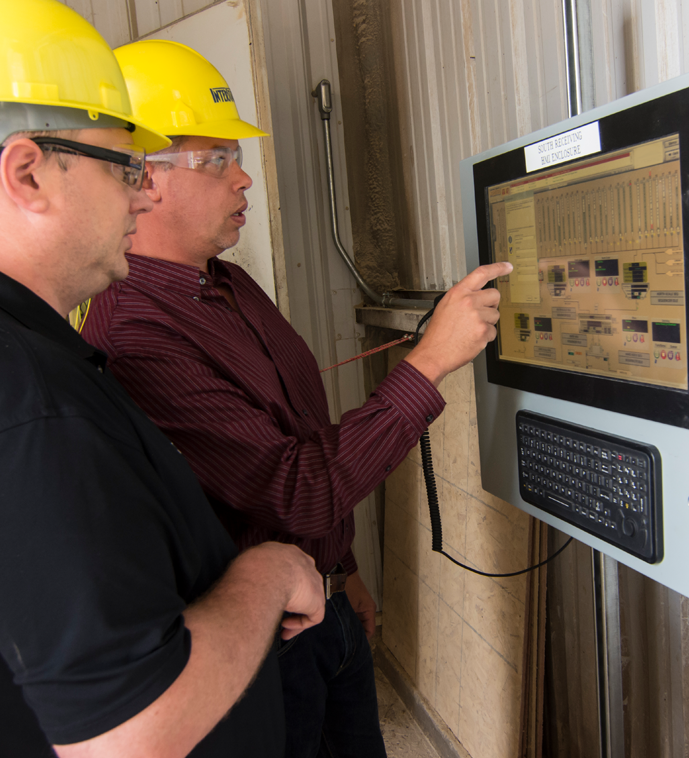 Two men wearing yellow hard hats and safety glasses are standing at an industrial touchscreen panel, with one of them pointing to data on the screen.