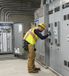 An electrician wearing a hard hat and high-visibility safety vest works inside an industrial electrical room.