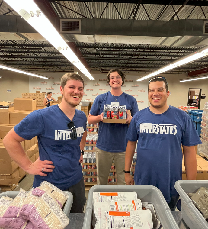 Three employees in Interstate branded t-shirts volunteering at food bank, smiling while organizing donation boxes and supplies