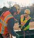 Two construction workers wearing safety gear work together outdoors on a job site. The worker in the foreground, wearing an orange Interstates safety vest and yellow hard hat, leans over while handling tools. The worker in the background, also in a yellow hard hat and bright safety vest, holds a cable and smiles.
