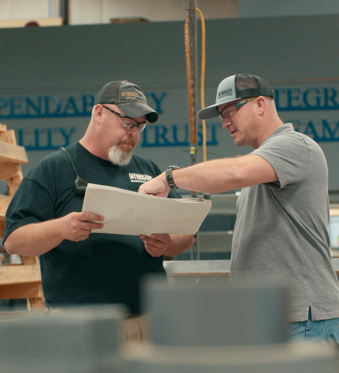 Two men wearing Interstates-branded hats and workwear stand in a fabrication area, closely reviewing a printed plan.