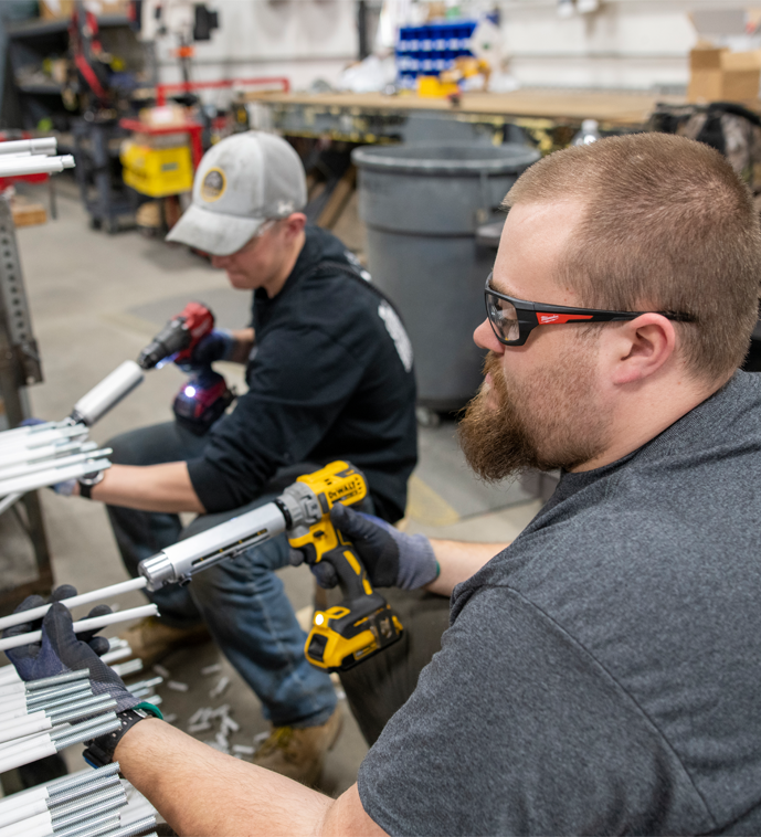 Two Interstates team members wear safety glasses and gloves as they use power tools to prepare conduit sections at a workstation in a fabrication shop.