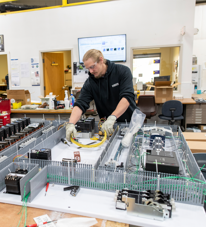 A technician wearing gloves and safety glasses works on a horizontal electrical control panel layout, carefully routing yellow and green cables in a bright, organized workspace.