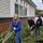 Three people working together outdoors beside a brick-and-siding building, using garden tools to clear bushes and leaves near a window.