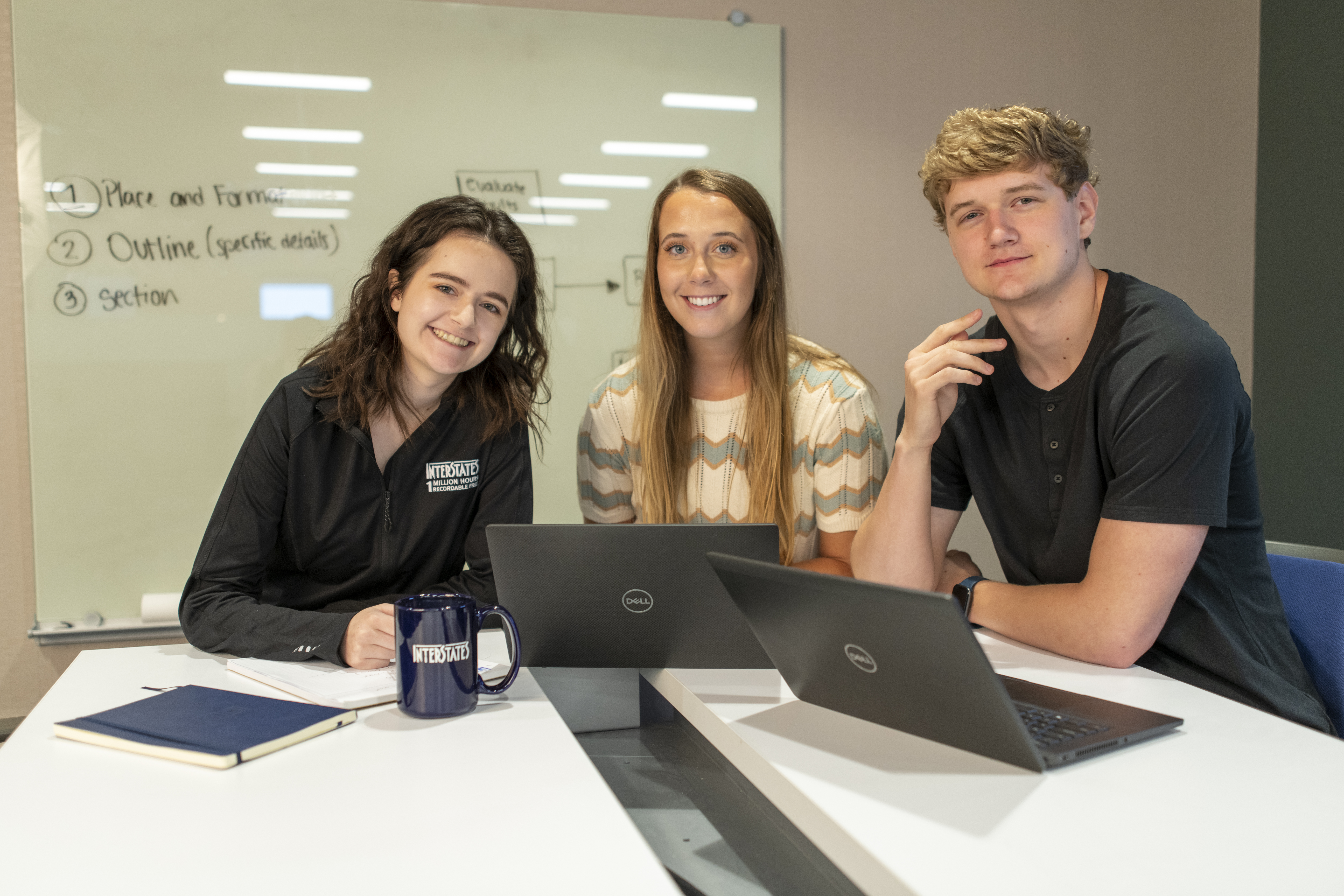 Three interns collaborating at table with laptops in meeting room, Interstate coffee mug visible, with whiteboard notes in background