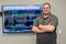 Technical designer in gray polo standing confidently in front of large display showing AutoCAD electrical schematics