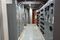 Narrow electrical room with rows of gray motor control center (MCC) panels lining both sides of a central walkway. Overhead lighting and conduit run along a wood-paneled ceiling, and the space features concrete walls and a metal-plated floor path.