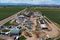 Aerial view of an industrial water treatment facility under construction, featuring multiple circular tanks, rectangular structures, construction equipment, and access roads, surrounded by green agricultural fields with mountains in the distance.