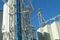 Tall grain facility structures under a clear blue sky, featuring a steel bucket elevator tower with a blue vertical conveyor, surrounded by large white storage silos.