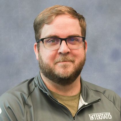 Portrait of a man with glasses and a short beard wearing a gray Interstates shirt, posed against a soft gray background.