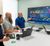 Three coworkers in a conference room review a large wall-mounted dashboard display showing colorful performance charts while seated around a table with laptops and notebooks.
