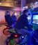 Three men sit on neon-lit motorcycle arcade games in a game room.