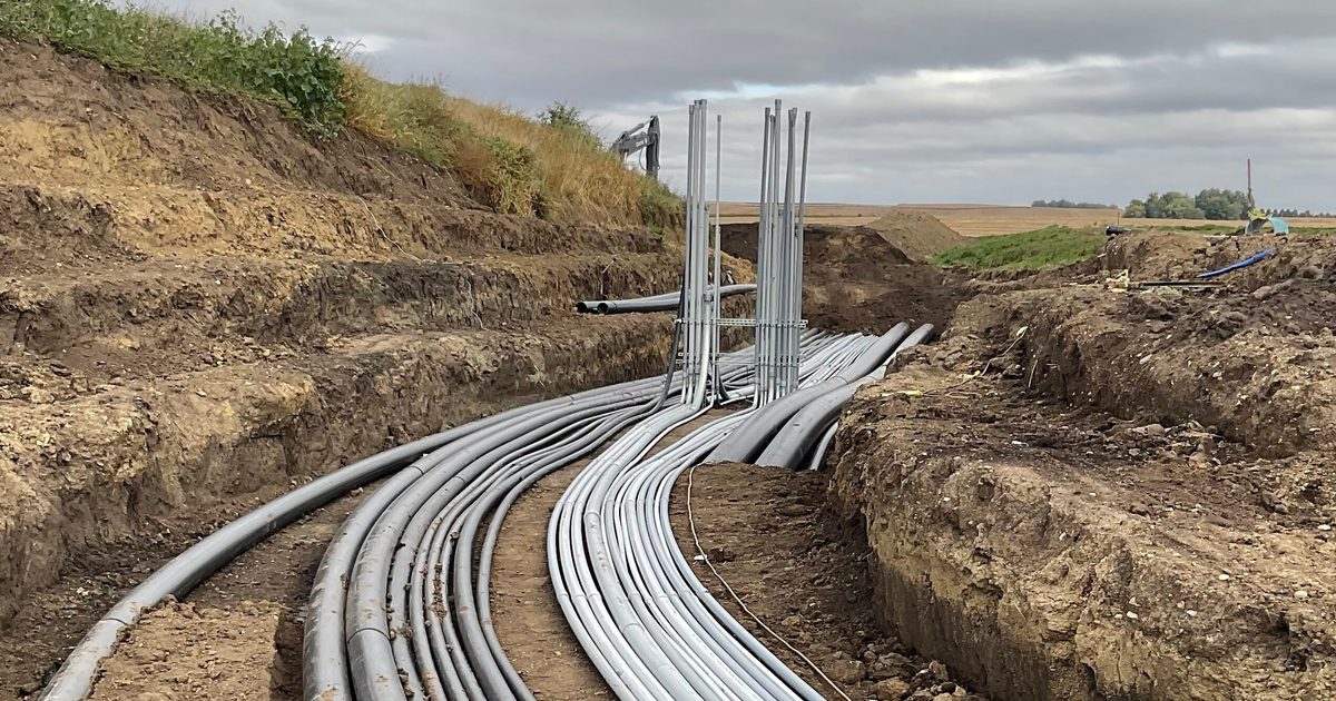 Underground electrical conduits run through a freshly dug trench at a construction site in a rural area, with multiple gray and black pipes curving toward a vertical conduit rack in the distance under a cloudy sky.