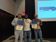 Three men stand onstage holding safety achievement certificates, with a presentation screen behind them displaying “Champion of Safety” and “FL Excellence.”