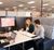 Three coworkers sit around a shared office table in a cubicle workspace, reviewing plans on a large wall-mounted monitor and laptop during a collaborative meeting.