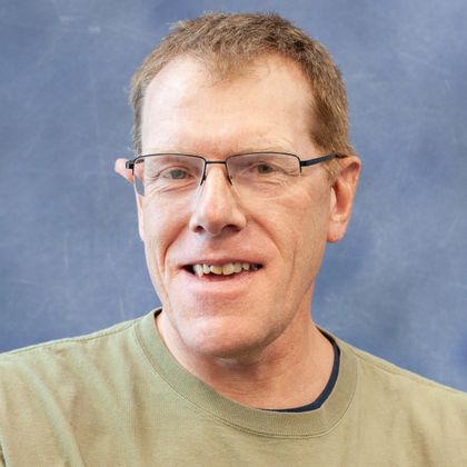 Close-up portrait of a man wearing glasses and a light green shirt, smiling against a blue background.