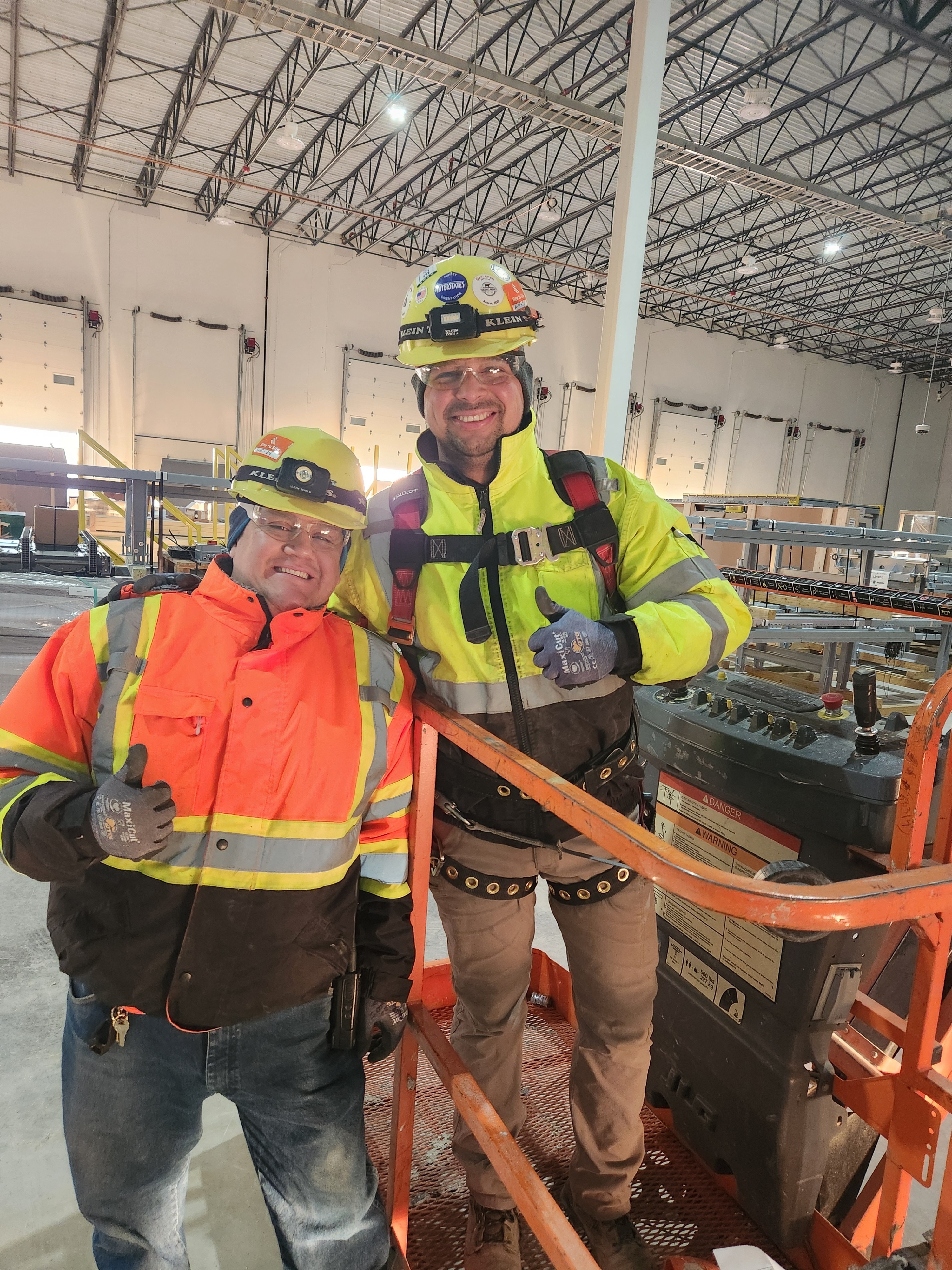 Two workers in hard hats and high-visibility gear giving thumbs up while working on industrial equipment in warehouse setting
