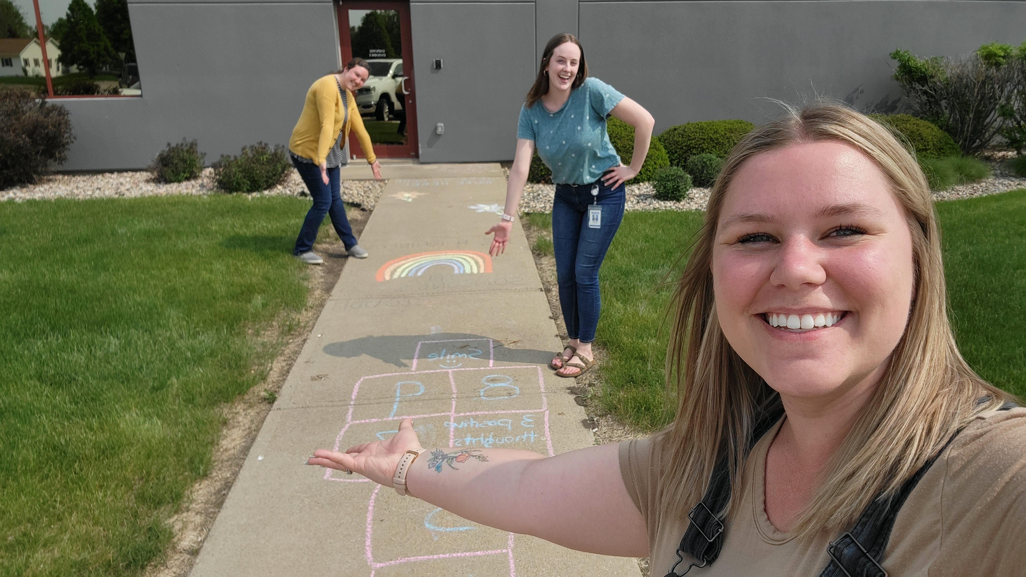 Colleagues showcase sidewalk art featuring a chalk hopscotch course and rainbow, taking selfie-style photo while gesturing to their creations