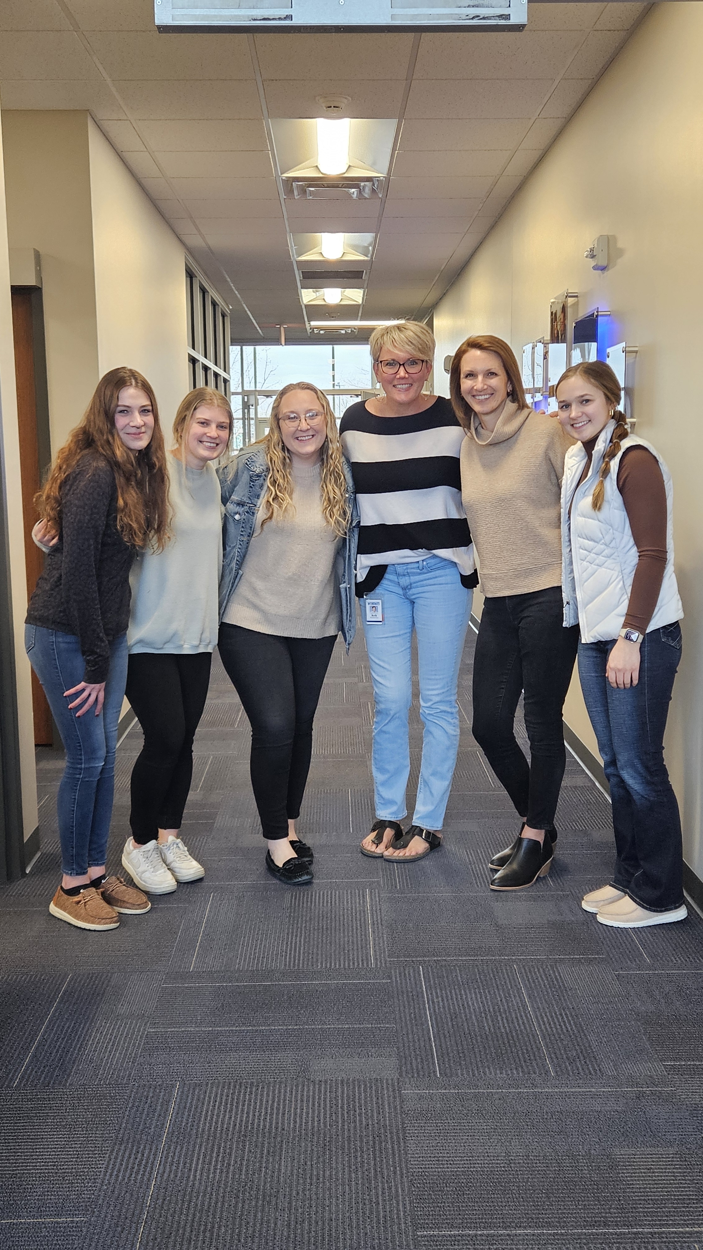 Six casual-dressed office workers standing together in a workplace hallway, wearing complementary neutral-toned outfits